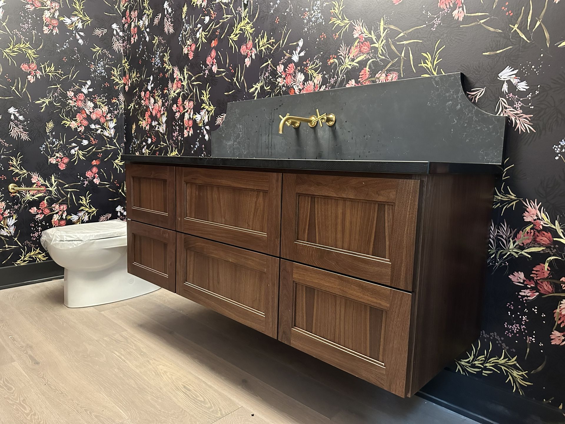 Modern bathroom vanity with dark wood cabinets, black countertop, and floral wallpaper in the background