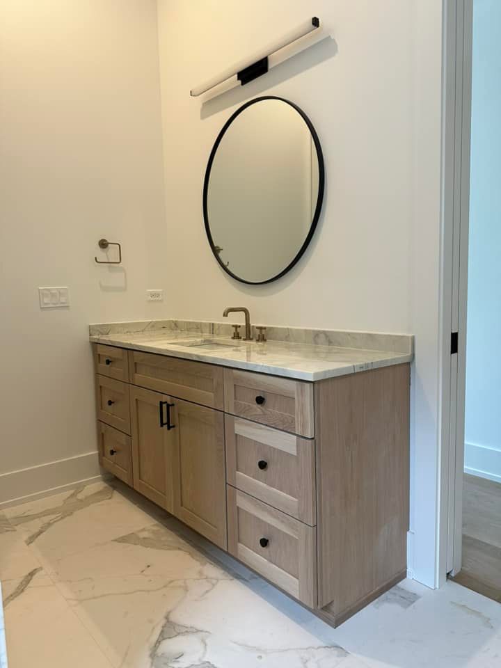 Modern bathroom vanity with marble countertop, round mirror, and wood cabinets in a bright, unfinished space