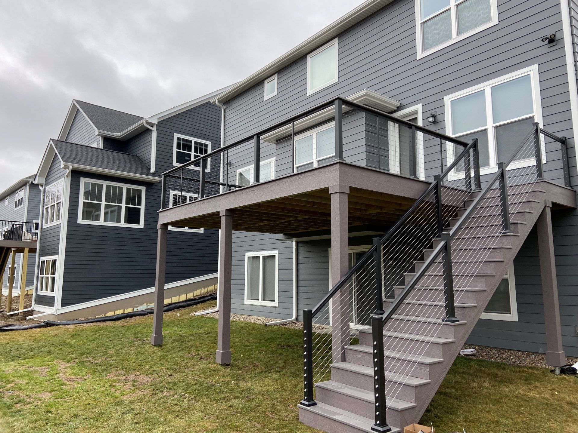 A modern house with dark gray siding features a raised composite deck with black railings and a staircase to the yard.