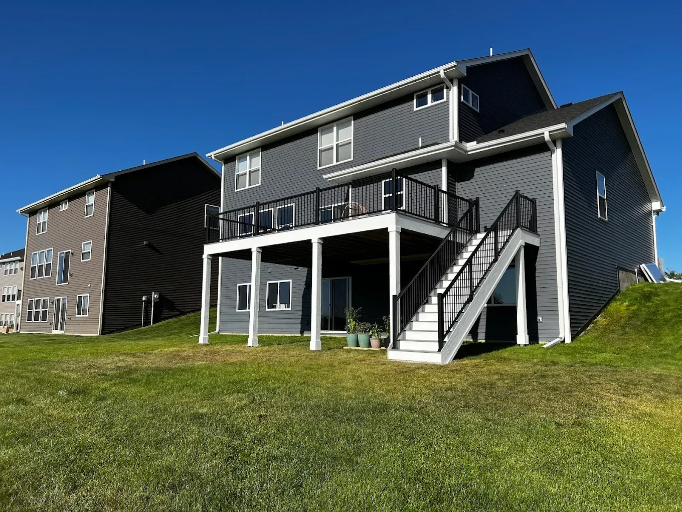 A dark grey two-story house with a wooden deck and white staircase, seen from the backyard on a sunny day.