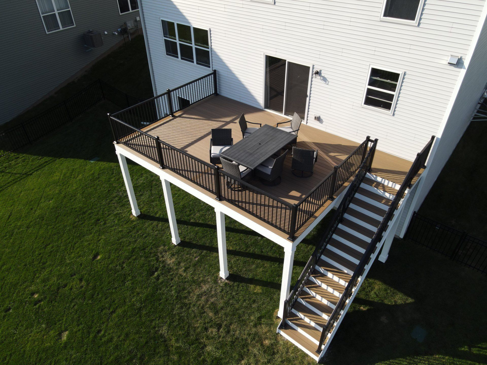 High-angle view of a tan elevated deck with a dining set and stairs leading down to a green lawn behind a white house.