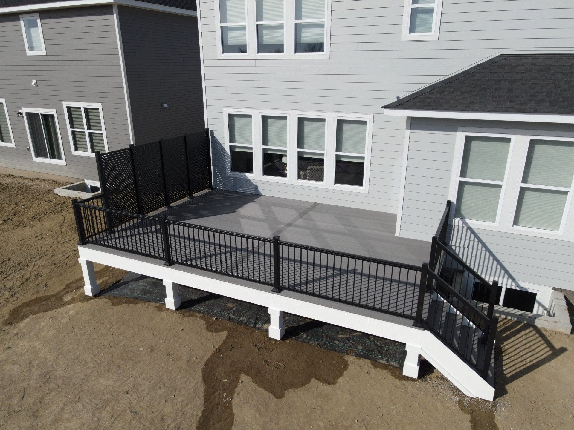 An elevated gray deck with black railings attached to the back of a light-gray suburban house with a dirt yard.