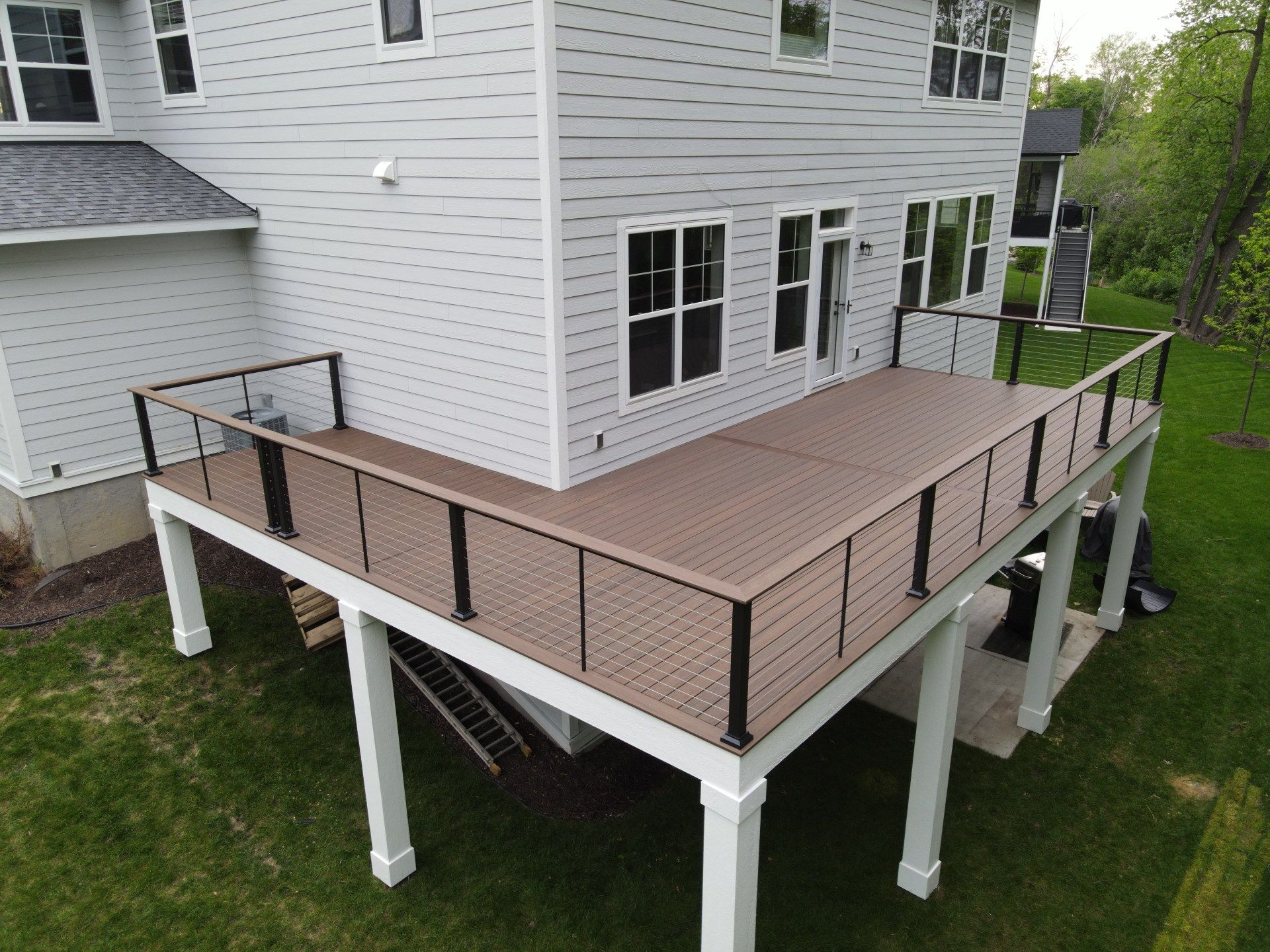 A gray house with a tan composite deck featuring black railings, supported by white posts, overlooking a green backyard.