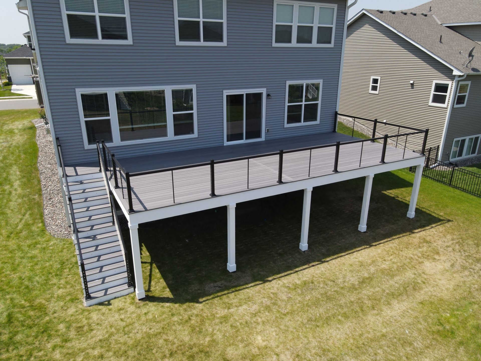 An elevated grey composite deck with black railings and stairs, attached to the back of a two-story blue house.