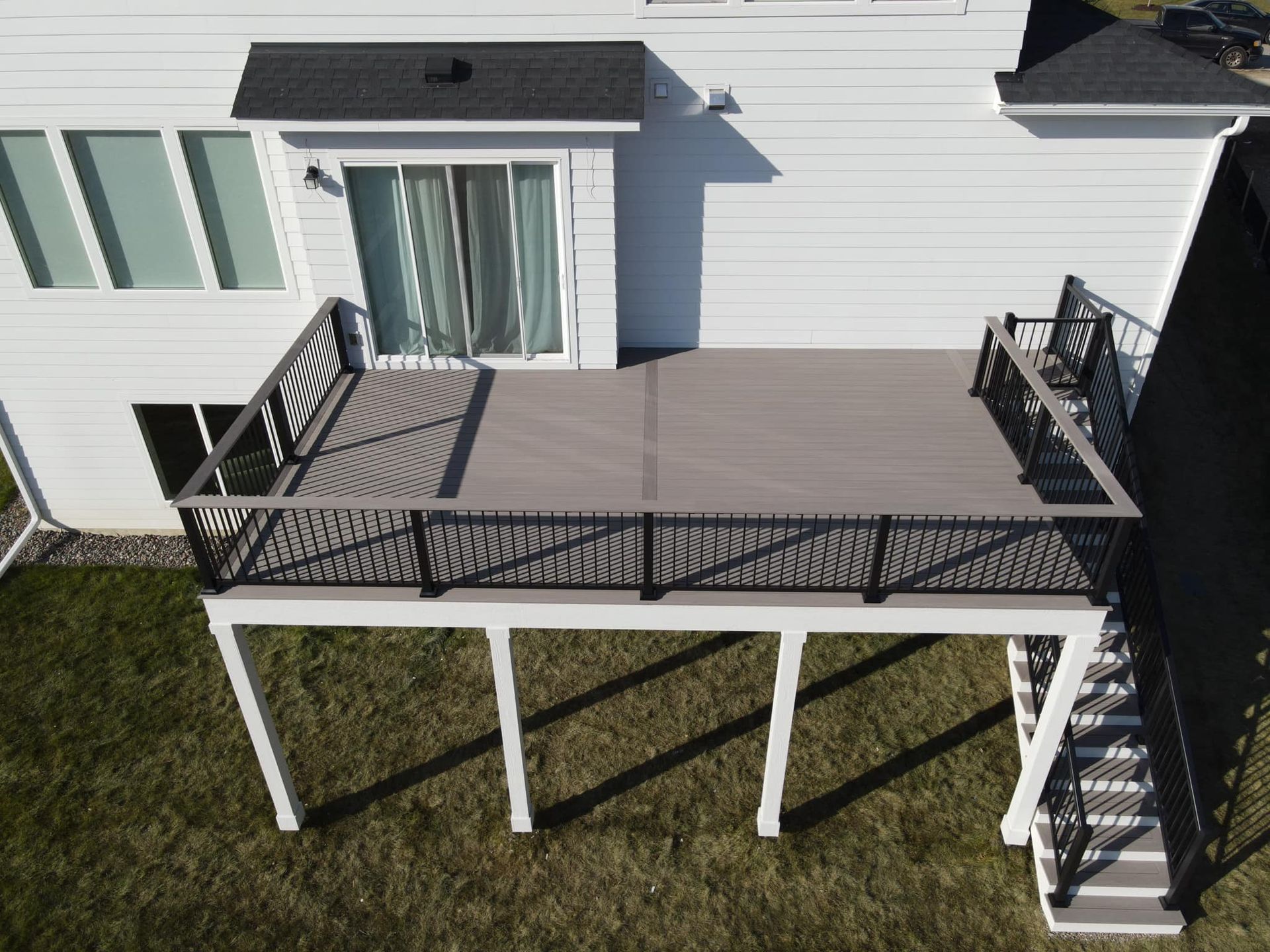 An aerial view of a white residential house with a gray composite deck, black railing, and stairs leading to the lawn.