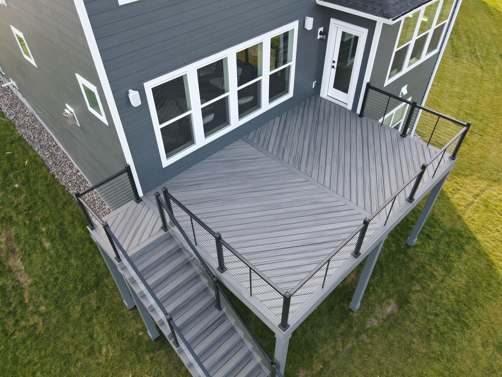 An elevated, gray composite deck with a diagonal pattern, black railing, and stairs leading down to a green lawn.