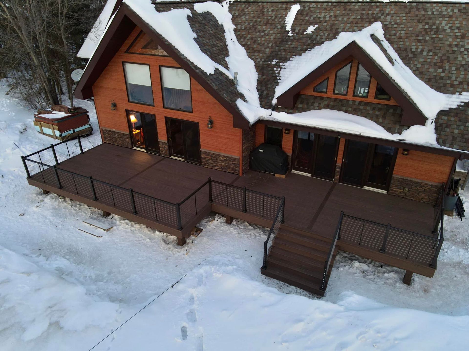 A two-story log cabin with a large, dark brown deck covered in snow, viewed from an elevated, side-angle perspective.