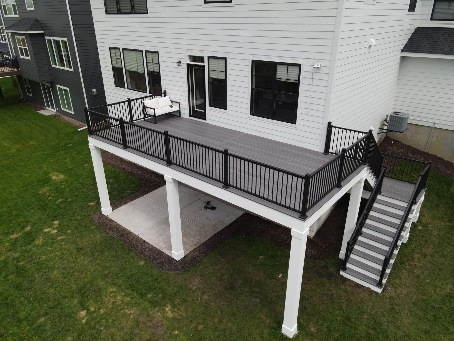 An elevated gray composite deck with black railings and stairs, attached to the back of a white house with a patio below.