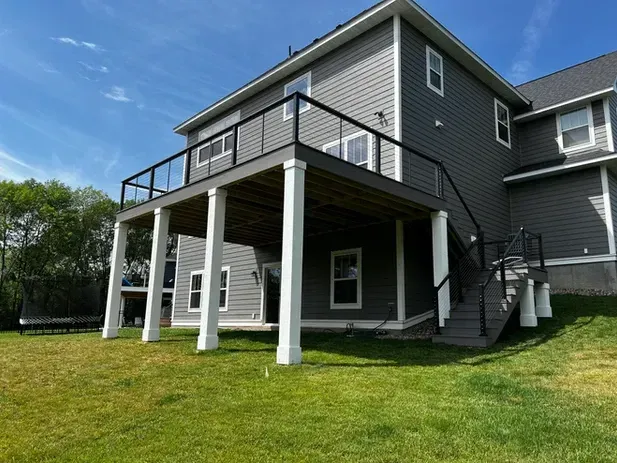 A grey two-story house with a large elevated deck featuring white support columns and a staircase leading to the yard.