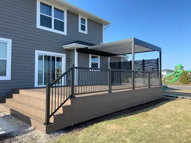 A multi-level deck with a metal pergola and railing attached to the back of a grey-sided house on a sunny day.