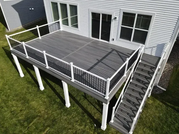 An elevated gray composite deck with white railings and stairs leading down to a green lawn attached to a white house.