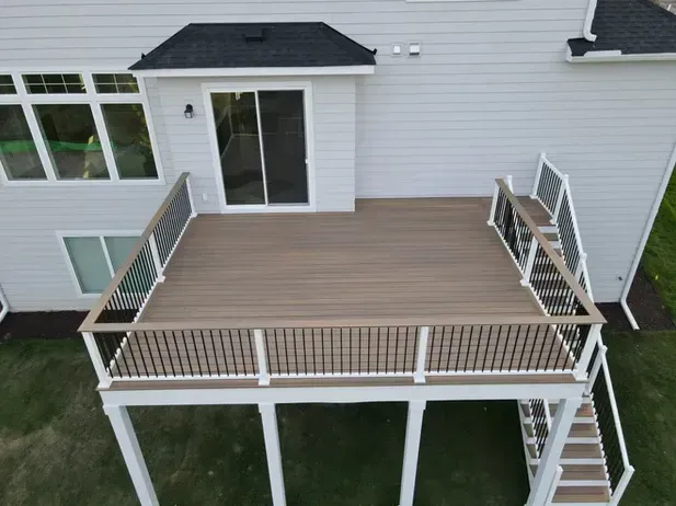 An elevated tan composite deck with white railings and stairs, attached to the back of a light gray two-story house.
