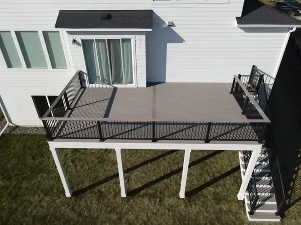 An elevated deck with gray composite flooring, black railings, and a staircase, attached to the back of a white house.
