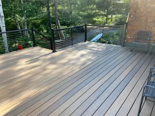 A wooden deck with a black cable railing overlooking a wooded area, with patio chairs on the right side.