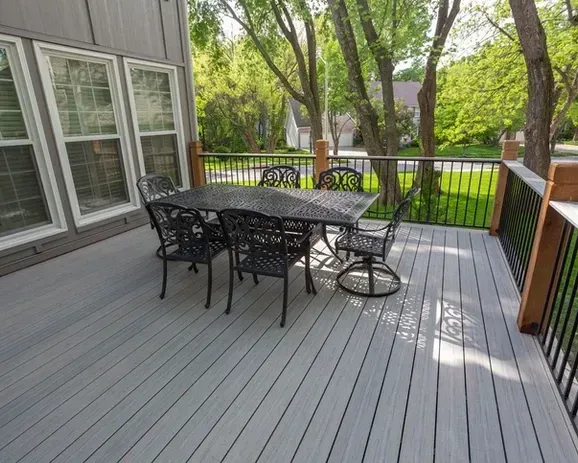 A patio dining set on a gray composite deck with black metal railings, overlooking a grassy yard with trees.
