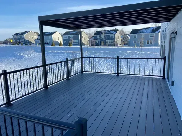 A modern gray deck with black metal railings and an overhead pergola, overlooking a snow-covered yard and neighborhood.