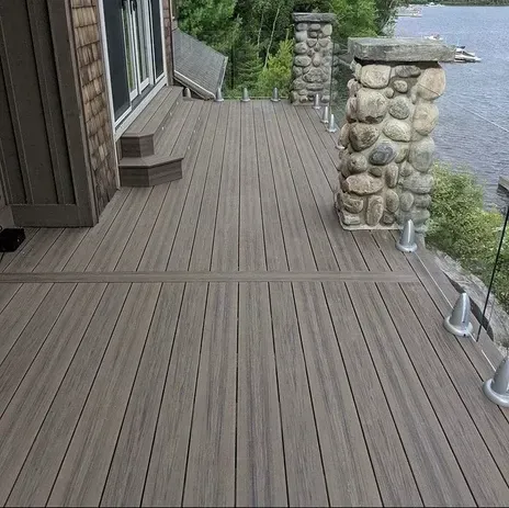 A gray composite deck with a stone pillar, a stepped entryway, and a glass railing overlooking a lake.
