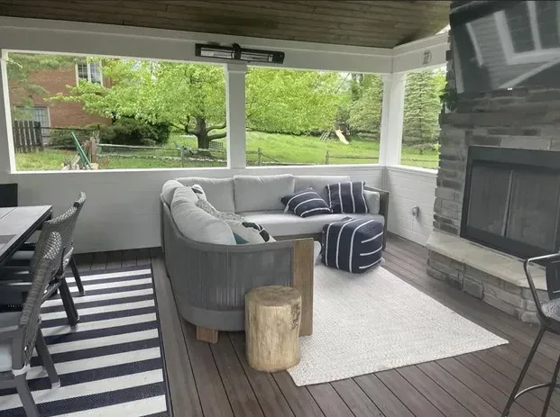 A covered outdoor patio featuring a gray sectional sofa, a striped ottoman, a fireplace, and dining seating on a deck.
