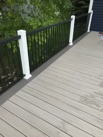 A gray composite deck featuring white railing posts and black vertical metal balusters against a leafy outdoor background.