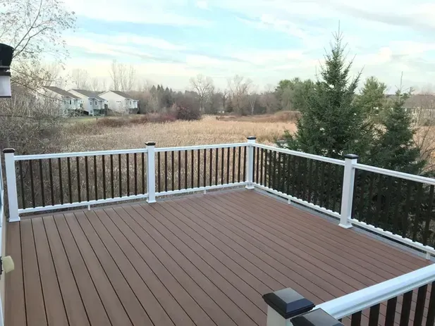 A brown composite deck with white railings overlooks a grassy field and trees under a cloudy sky.