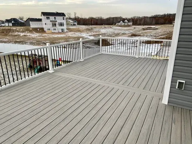 A gray composite deck with a white railing overlooks a snowy, open field with houses in the distance.