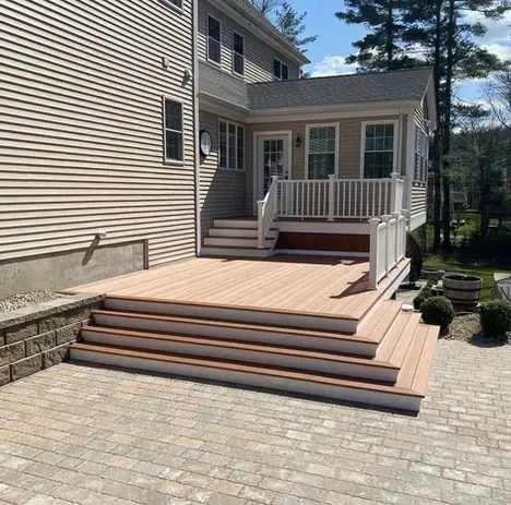 A wide wooden deck with white railings and broad steps leading down to a stone patio next to a two-story beige house.