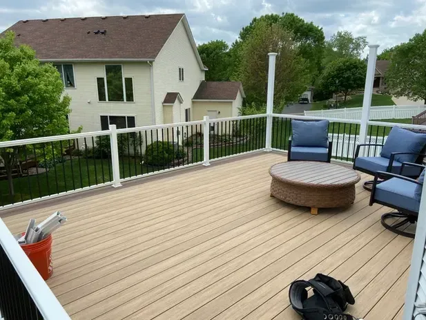 A beige deck with a round woven table, two blue chairs, and an orange bucket overlooking a yard and suburban homes.