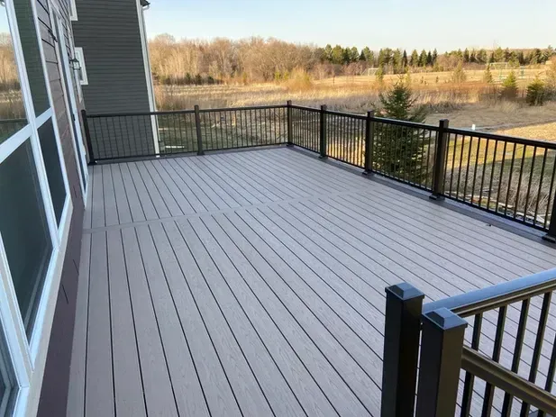 A gray composite deck with black metal railings overlooking a grassy field and trees on a sunny day.