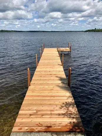 A new, light-colored wooden dock extends into a calm lake under a cloudy blue sky.
