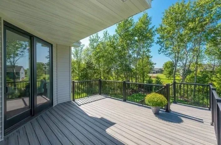 A gray deck with black railings overlooks a lush green yard surrounded by tall trees under a bright blue sky.