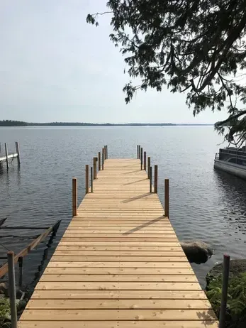 A newly constructed wooden dock extends into a calm lake under a hazy sky, with trees visible overhead.