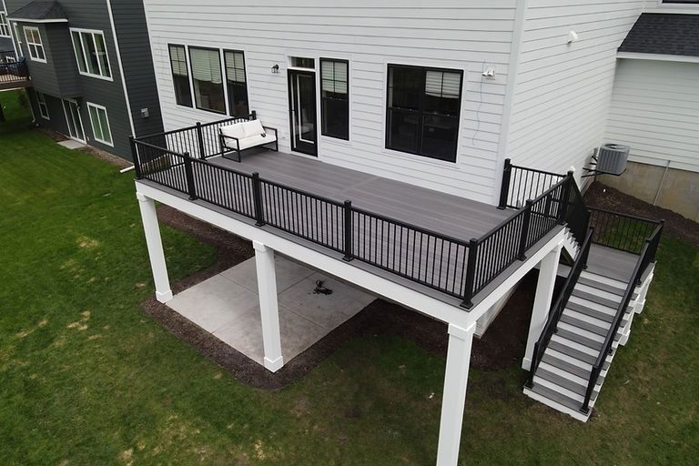 An elevated grey composite deck with black railings and stairs, positioned above a patio on a suburban white house.