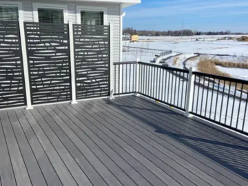 A grey composite deck with black metal railing and decorative privacy screens overlooks a snowy landscape.