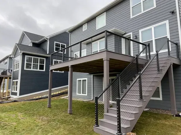 A gray-sided house with a raised wooden deck featuring cable railings and stairs leading down to a grass lawn.