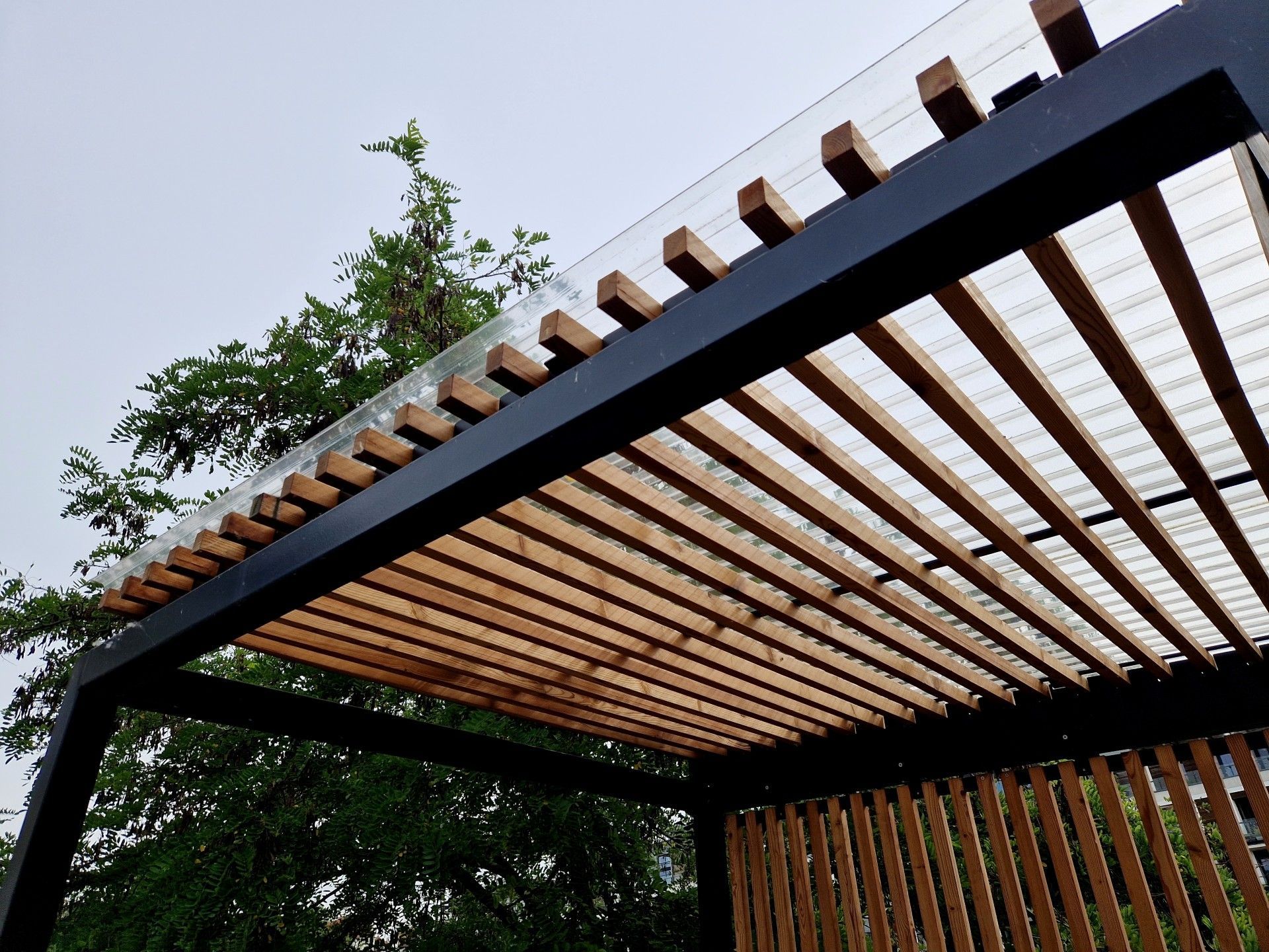 A modern outdoor pergola with a black metal frame and light-colored wooden slats against a cloudy sky.