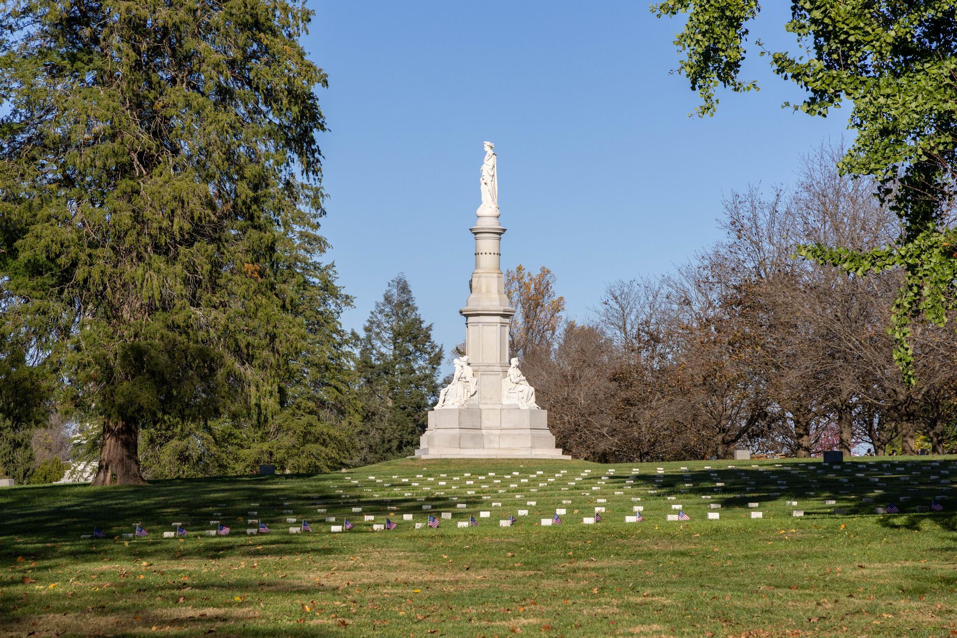 White monument on a grassy hill in a park, framed by trees under a clear blue sky.
