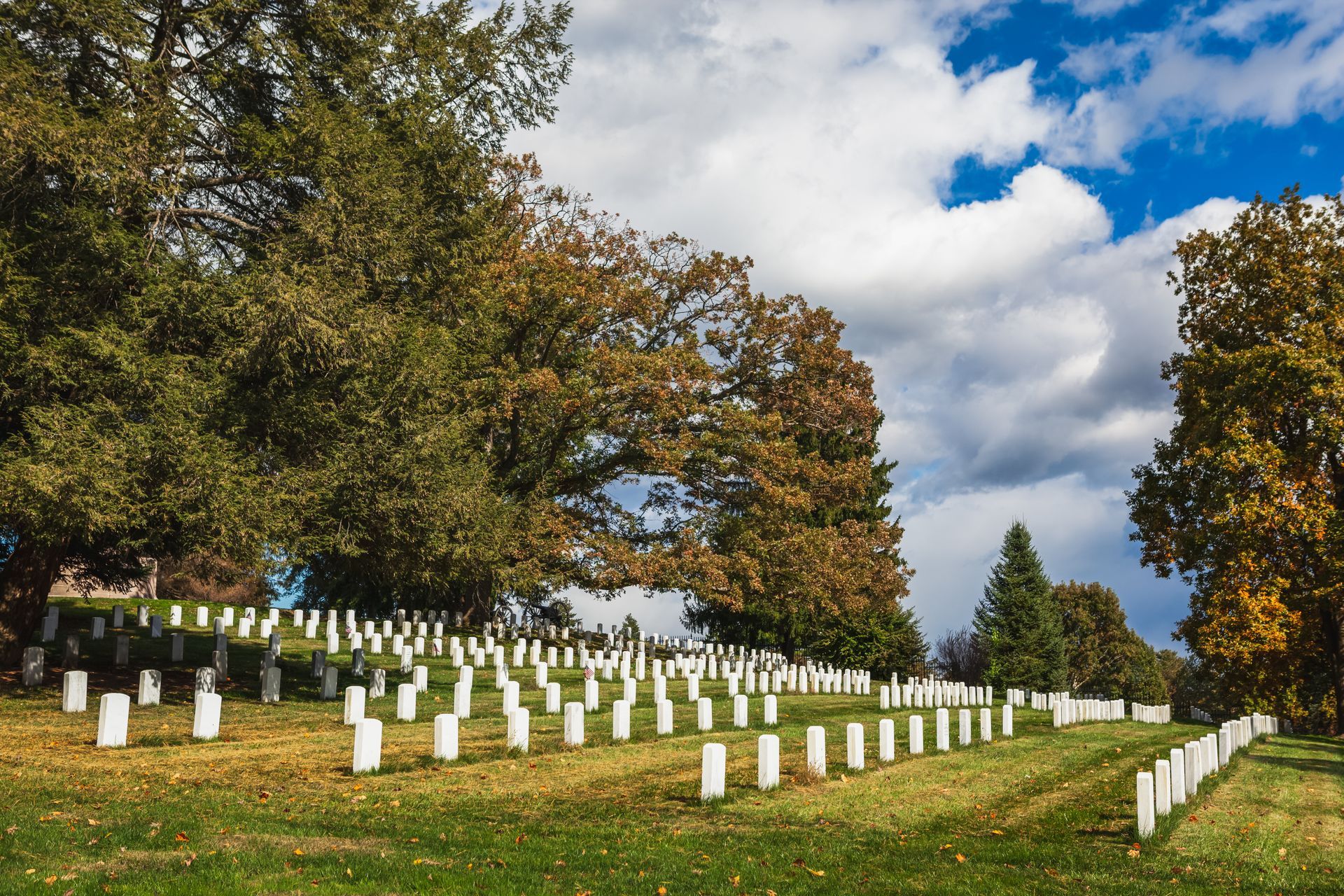 Row of white headstones in a cemetery beneath autumn trees and a cloudy sky
