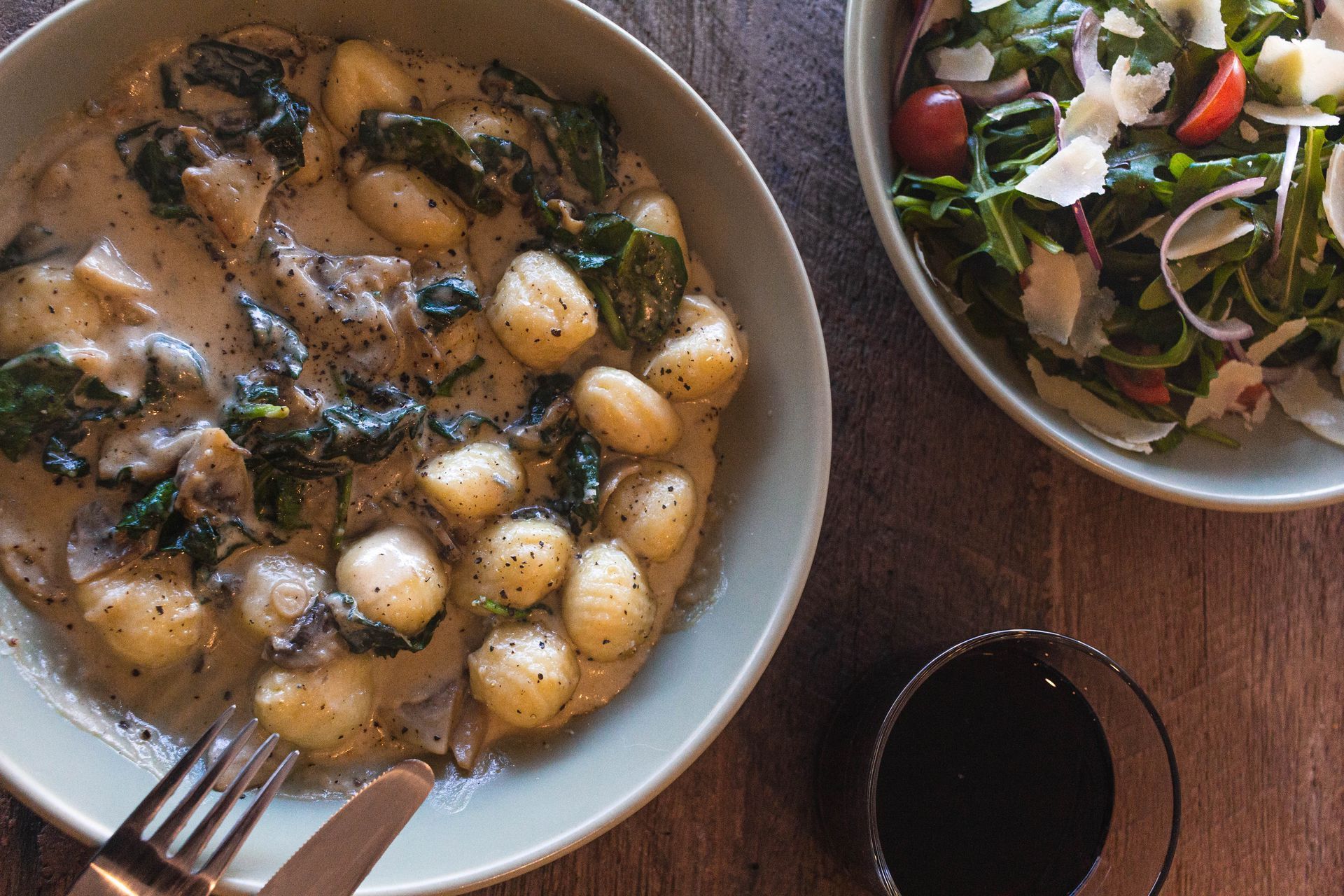 Plate of gnocchi with brown sauce and herbs beside a fresh green salad on a wooden table