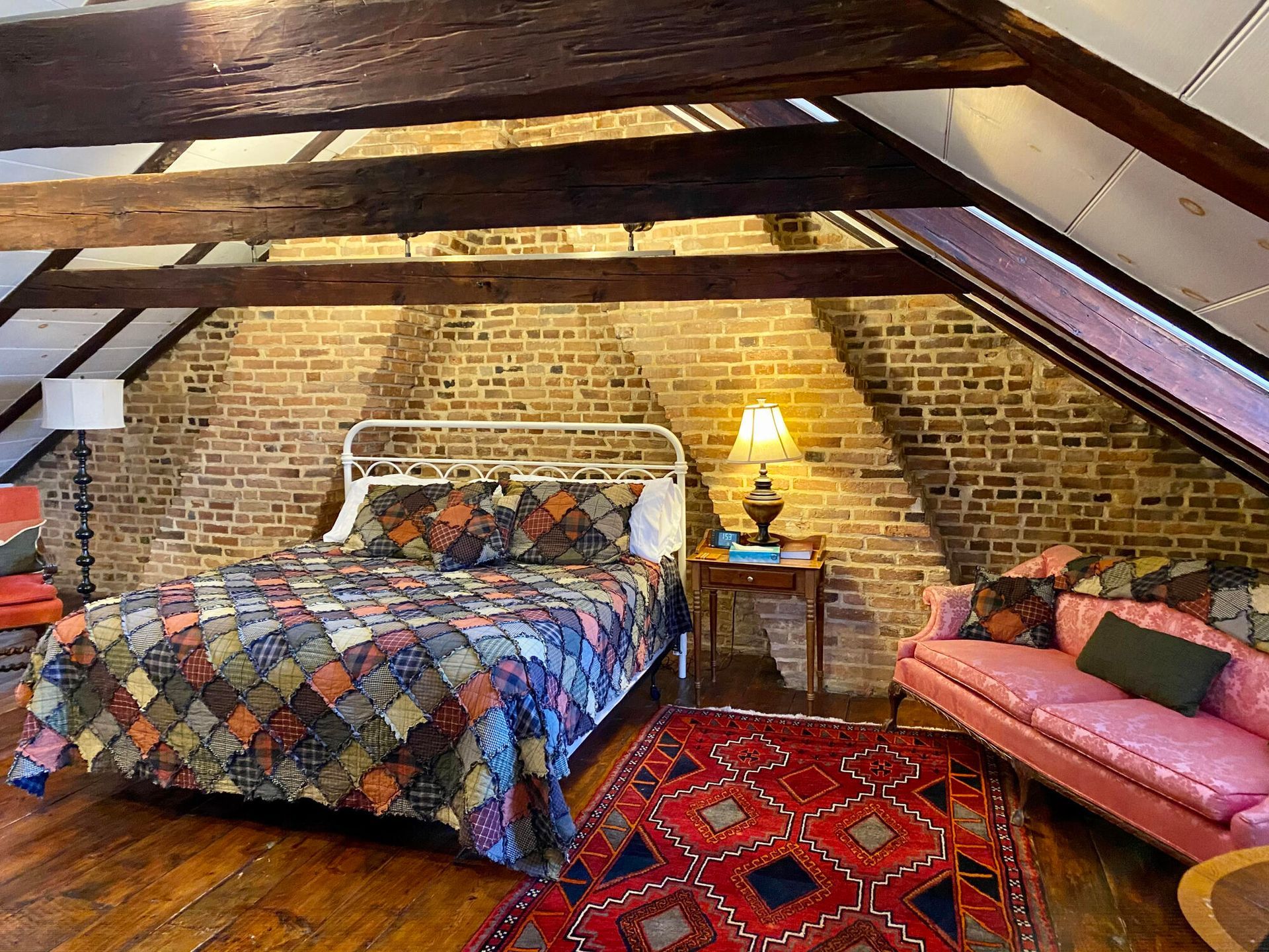 Cozy attic bedroom with brick walls, colorful quilted bed, red rug, and pink sofa under sloped beams