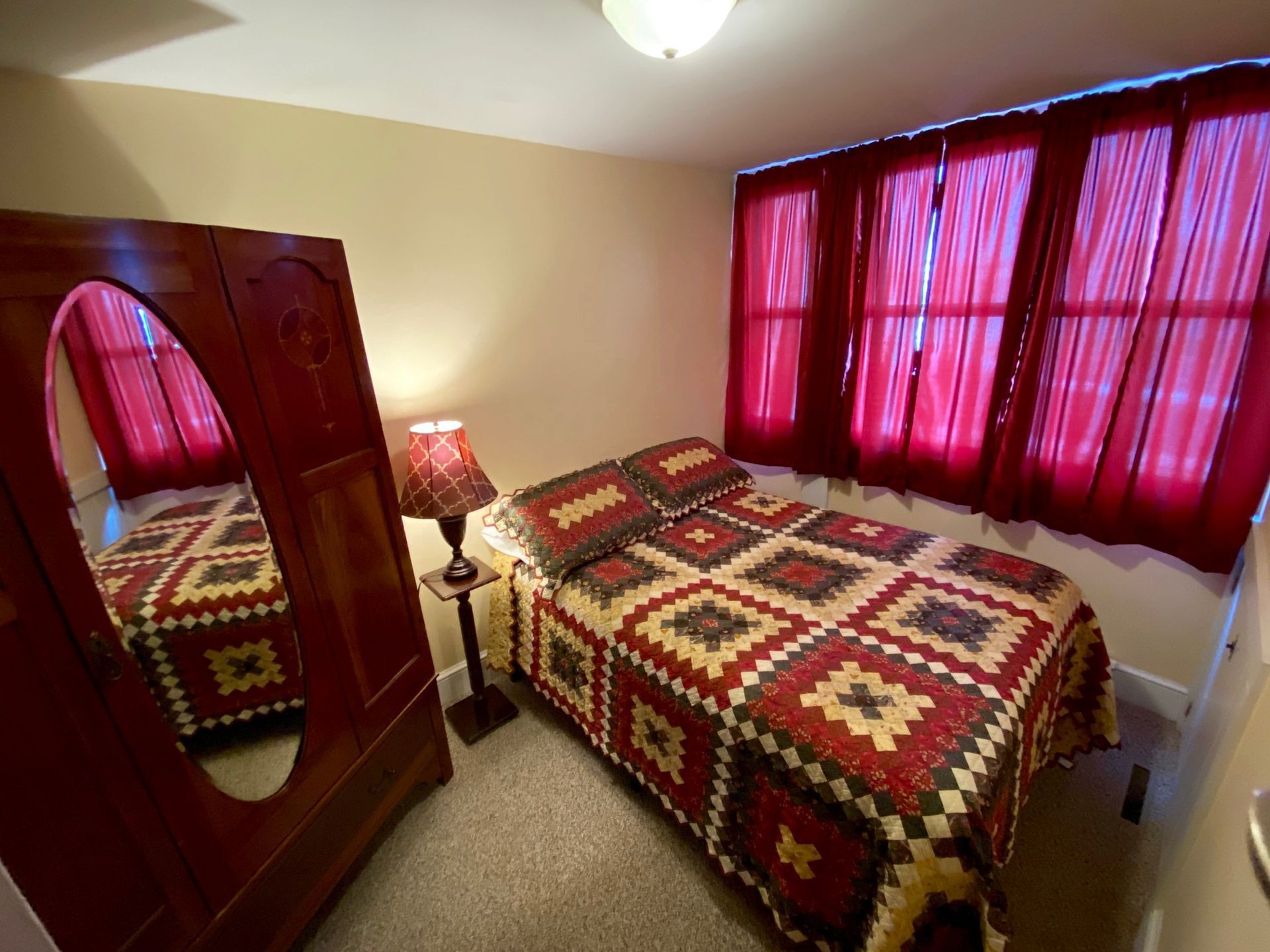 Bedroom with two beds, red patterned quilts, matching red curtains, and a wooden wardrobe with a mirror.