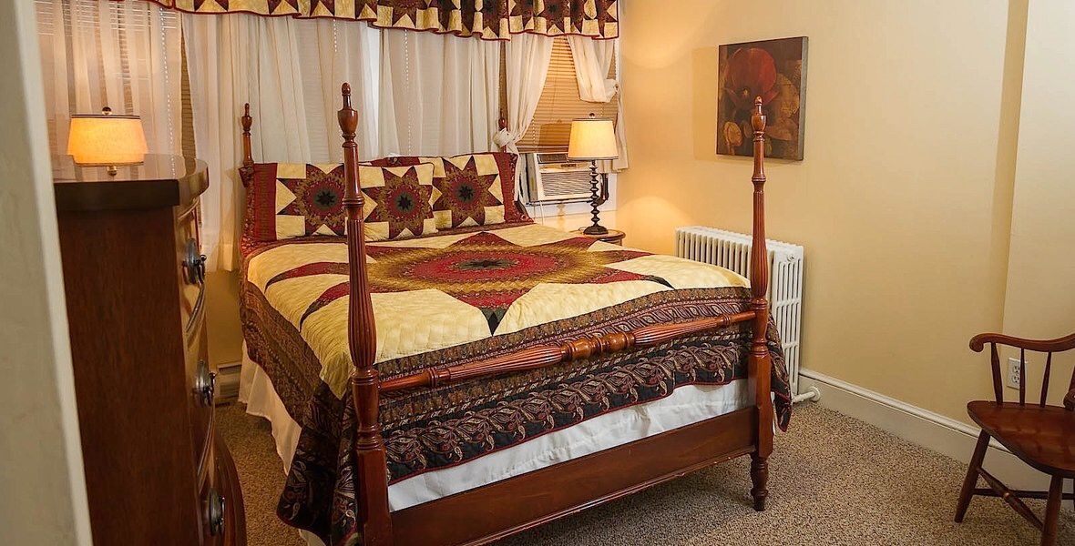 Cozy bedroom with a bed, patterned quilt, bedside lamps, and a chair near the window.