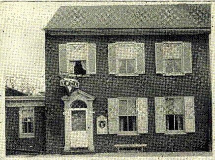 Old black-and-white photo of a two-story brick house with a storefront-style entrance and upper windows.