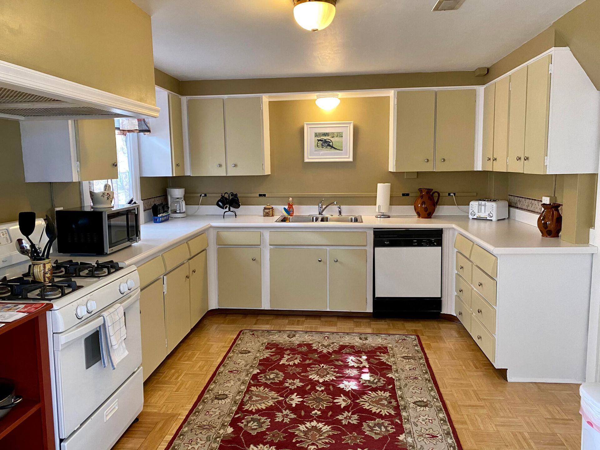 Small beige kitchen with white cabinets, wood floor, and a red patterned rug.