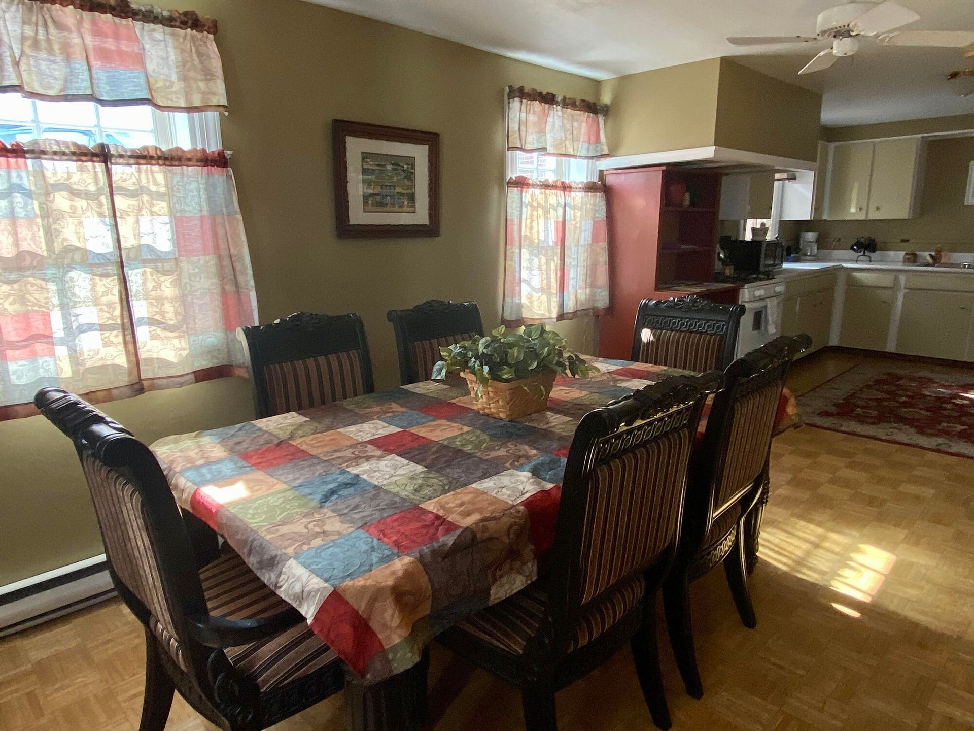 Dining room with checkered tablecloth, dark chairs, and sunlight through curtains, open to a kitchen area