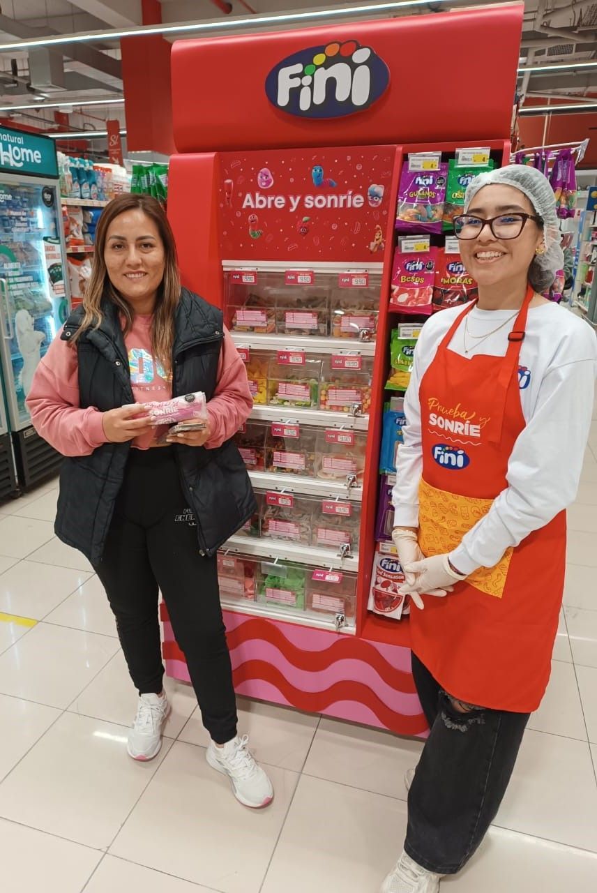 Dos personas posan junto a un expositor rojo de caramelos Fini en el pasillo de un supermercado; ambas sonríen.