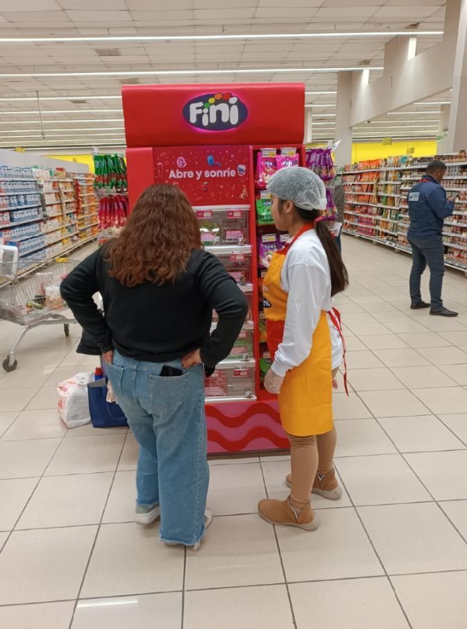 Una empleada de la tienda, con un delantal naranja, ayuda a una clienta junto a un colorido expositor de caramelos