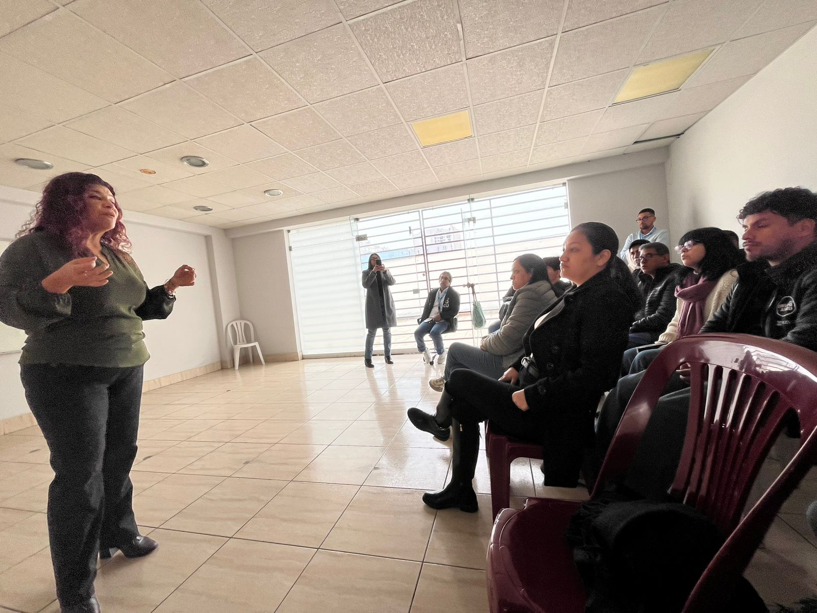 Una persona se pone de pie y habla a un grupo sentado en sillas en una habitación luminosa