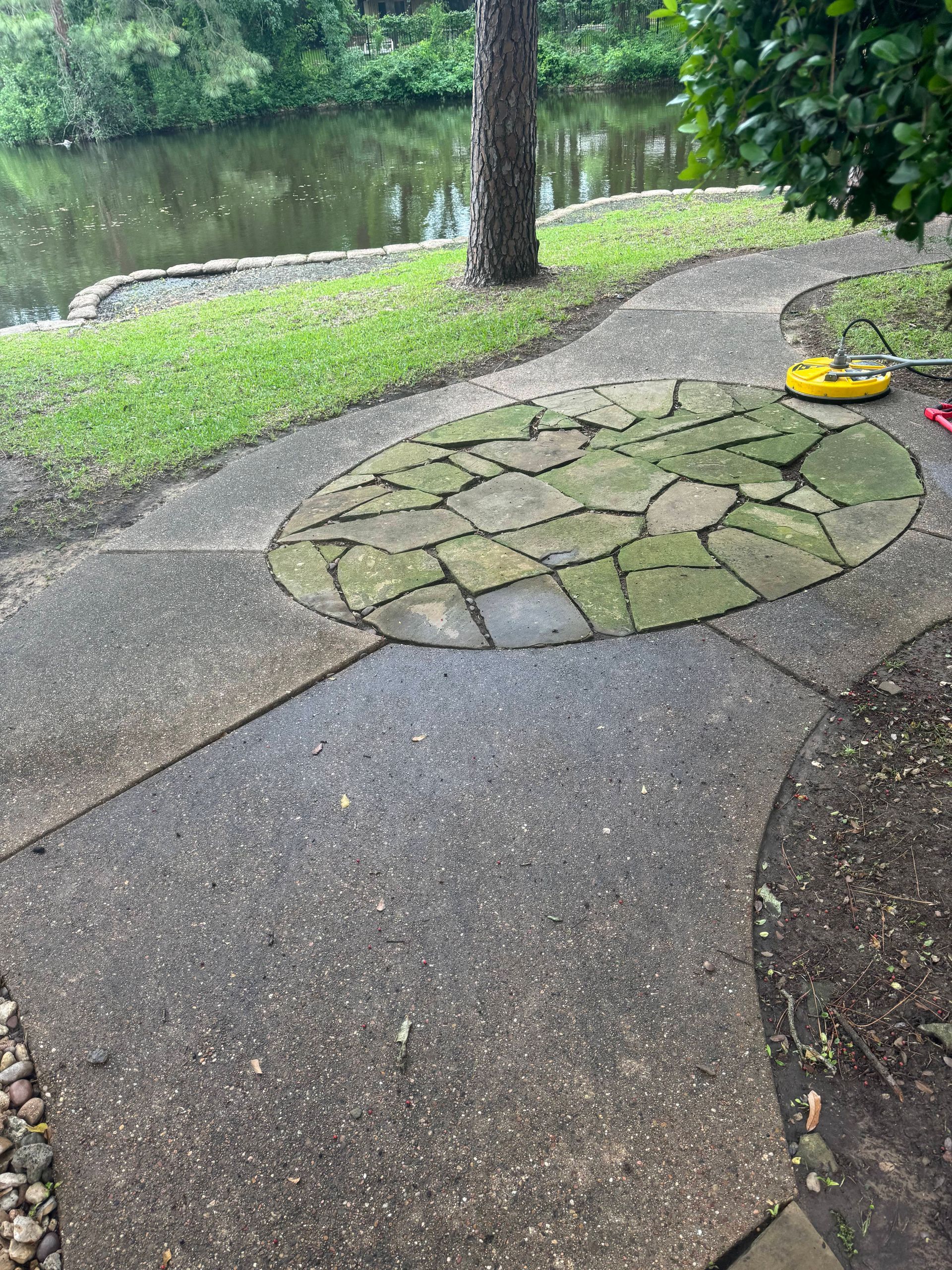 A concrete walkway with a stone circle in the middle and a pond in the background.