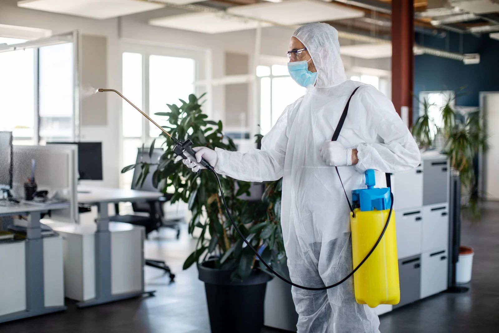 Person in hazmat suit sprays disinfectant in an office with desks and plants.