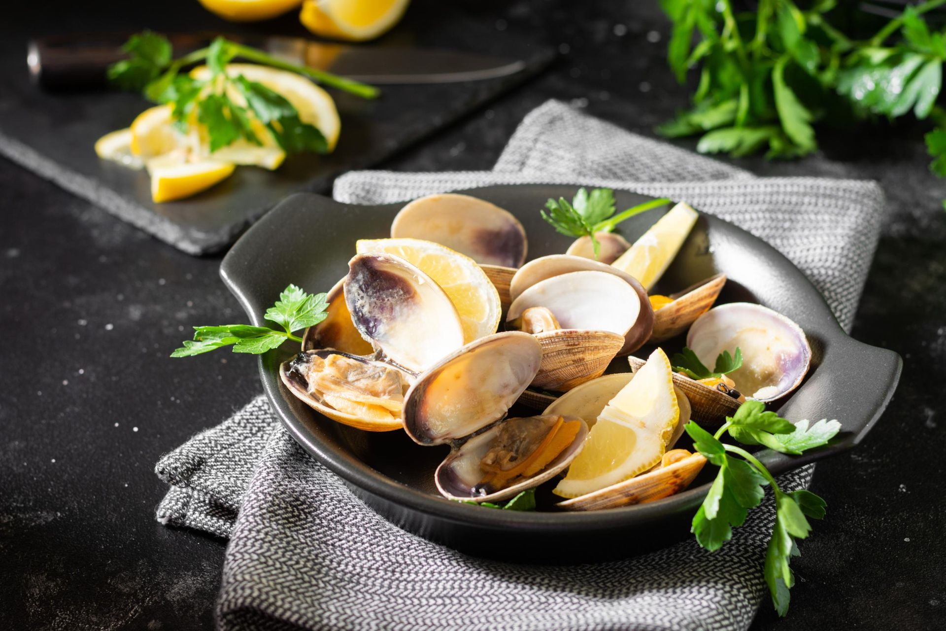 A bowl of steamed clams garnished with parsley and lemon wedges, set on a gray cloth against a dark surface.